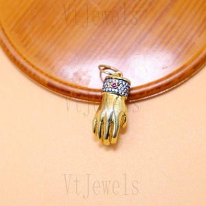 Op de afbeelding: Een goudkleurige handhanger met een ring van kleine witte en rode stenen rond de pols. De hanger is opgehangen aan een kleine gouden lus. De hand heeft de vorm van een vuist.