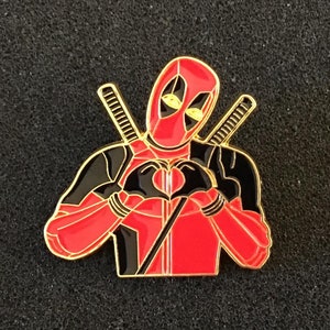 Op de afbeelding: Een rode en zwarte emaille pin met Deadpool die een hartvorm maakt met zijn handen. De pin heeft een gouden afwerking en is geplaatst tegen een zwarte achtergrond.