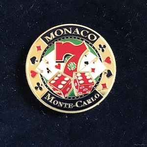 May include: A circular, gold-coloured casino token featuring the words "MONACO" and "Monte-Carlo". The design includes a red seven, dice, and playing card suits on a black background.