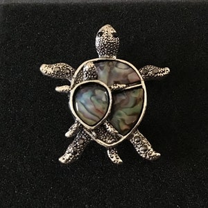 Puede incluir: Un broche de tortuga de plata con una incrustación de concha de abulón iridiscente. La concha tiene un patrón de color verde, azul y morado.
