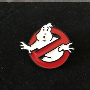 May include: A red and white enamel pin featuring the Ghostbusters No Ghost logo. The logo is a red circle with a diagonal line through it. A white ghost with a surprised expression is inside the circle.