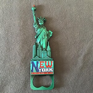 May include: Green metal bottle opener featuring a depiction of the Statue of Liberty and the words "NEW YORK" in red, white, and blue.