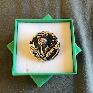 May include: A round brooch with a thistle design in a green box. The brooch has a gold-colored metal frame and a dark blue center. The thistle is detailed with gold-colored accents. The box is square with a white interior.