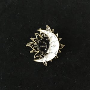Può includere: Uno spillo in smalto dorato con un sole nero e una luna crescente bianca con gli occhi chiusi. Il sole e la luna sono intrecciati, con la luna che copre parzialmente il sole.