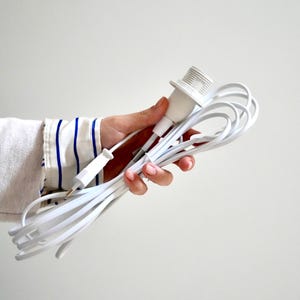 Könnte beinhalten: Ein weißer Lampenhalter mit einem weißen Kabel und einem weißen Stecker. Das Kabel ist aufgerollt und wird in einer Hand gehalten. Der Ärmel eines weißen Hemdes mit blauen Streifen ist sichtbar.