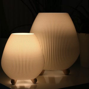 Könnte beinhalten: Zwei weiße Keramiklampen mit gerippten Schirmen und Holzbeinen. Die größere Lampe befindet sich im Hintergrund und die kleinere Lampe im Vordergrund.