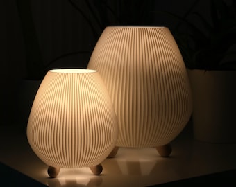 May include: Two cream-colored, teardrop-shaped lamps with vertical ribbed detailing. The lamps are illuminated from within, casting a warm glow. Each lamp rests on three small, rounded wooden feet. The background is dark, with hints of greenery.