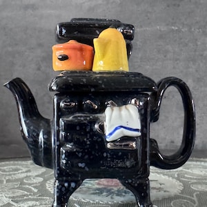 May include: A black ceramic teapot in the shape of a vintage stove, with a speckled finish. It has a spout, handle, and small decorative pots in orange and yellow. A small white towel with blue trim is on the front.