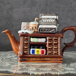 Könnte beinhalten: Eine braune Keramik-Teekanne in Form eines Ladens mit Registrierkasse, Schubladen und Gläsern. Die Teekanne hat einen gebogenen Ausguss und Griff, mit Details wie einem Teeglas und kleinen Boxen. Das Design ist aufwendig und dekorativ.