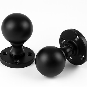 May include: Two black door knobs with a spherical design and a circular base. The knobs are made of a matte black material and are set against a white background. The base has four screw holes.