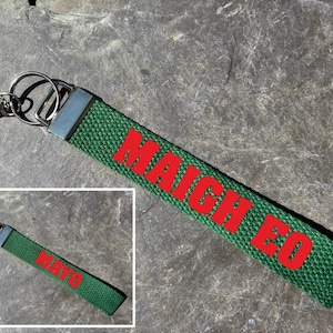 May include: Green fabric keychain with the red text "MAIGH EO". The keychain has a silver metal clasp and ring. The background is a textured, grey stone surface.