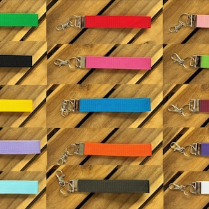Puede incluir: Una colección de llaveros de muñeca coloridos en varios colores, incluyendo verde, rojo, rosa, negro, amarillo, azul, morado, naranja, morado oscuro, turquesa y blanco. Cada llavero tiene una correa de tela y un cierre plateado.