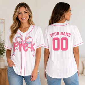May include: A white baseball jersey with thin pink pinstripes and pink trim. The front features a colourful rainbow bow graphic above the pink text "pre-k". The back displays the pink text "YOUR NAME" above the large pink number "00".
