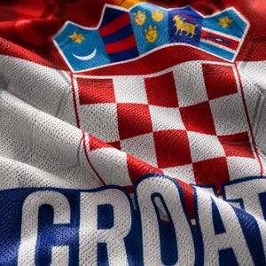 Pu&ograve; includere: Una maglia sportiva a scacchi rossi e bianchi con lo stemma croato nella parte superiore, che include una corona con cinque piccoli scudi. La parola CROATIA &egrave; stampata in grandi lettere maiuscole bianche su una fascia orizzontale blu.