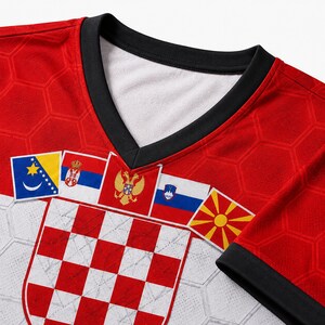 May include: A red and white V-neck jersey featuring a repeating hexagonal pattern. The front displays a large red and white checkered shield emblem below a row of five distinct national flags, including those of Bosnia, Serbia, Montenegro, Slovenia, and North Macedonia.