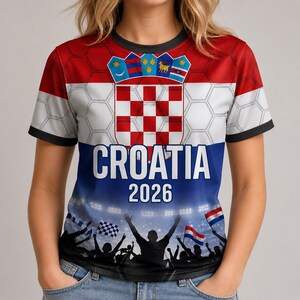 Pu&ograve; includere: Una t-shirt a maniche corte con i colori rosso, bianco e blu della bandiera croata, lo stemma della Croazia e una grafica di una folla in festa. Il testo CROATIA e 2026 &egrave; stampato in bianco su un motivo esagonale.
