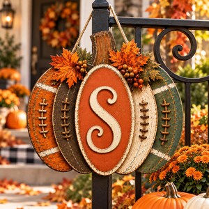 May include: A pumpkin-shaped wall hanging featuring a central oval with the letter S, flanked by segments resembling footballs with laces. The piece is decorated with artificial orange maple leaves and small berries, suspended by a jute rope.