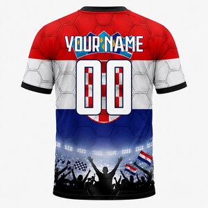 Pu&ograve; includere: Una maglia a maniche corte con un motivo della bandiera croata rossa, bianca e blu con una sovrapposizione esagonale. Il retro mostra il testo YOUR NAME sopra il numero 00, con una grafica di una folla da stadio sull'orlo inferiore.
