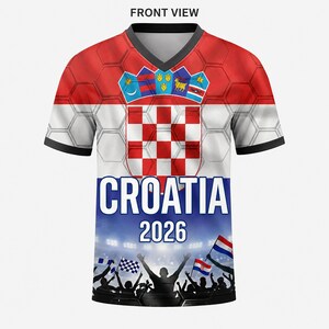 May include: A V-neck t-shirt featuring the Croatian flag with a red and white checkerboard pattern, the text CROATIA 2026, and a graphic of a cheering crowd with flags at the bottom. The design includes a honeycomb overlay and a coat of arms.