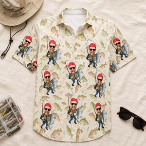 May include: A short-sleeved button-down shirt in a light beige colour featuring a repeating pattern of a cartoon fisherman wearing a red beanie and sunglasses while holding a fish, interspersed with various fish illustrations across the fabric.