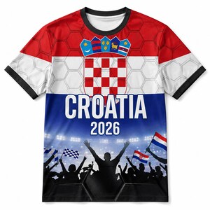 Pu&ograve; includere: Una t-shirt a maniche corte con un design rosso, bianco e blu che presenta lo stemma croato, il testo CROATIA 2026 e la grafica di una folla in festa sotto le luci dello stadio. Lo sfondo include un sottile motivo esagonale bianco.