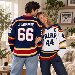 May include: A dark blue hockey jersey with the name DI LAURENTIS and number 66 in white and red, paired with a white hockey jersey featuring the text BRIAR, the number 44, and a letter C in dark blue, both with yellow and red sleeve stripes.