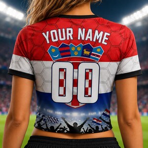 May include: A short-sleeved jersey featuring a red, white, and blue color scheme with a honeycomb pattern. The back displays the text YOUR NAME above a crest with five shields and the large white number 00. The bottom shows a stadium crowd graphic.