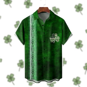 May include: Green short-sleeved shirt with a shamrock design. The shirt has a white and green Celtic pattern on the left side and a shamrock with an American flag design on the chest pocket. The shirt is hanging on a wooden hanger.