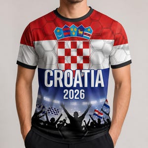 Pu&ograve; includere: Una t-shirt a maniche corte con la bandiera croata rossa, bianca e blu con un motivo a nido d'ape sovrapposto. Il design include lo stemma croato, la scritta "CROATIA 2026" in bianco e una grafica di una folla festante in uno stadio.