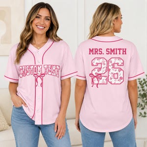 May include: A light pink button-down baseball jersey with dark pink piping. The front features the text CUSTOM TEXT with a bow graphic. The back displays the text MRS. SMITH above the number 26, both featuring a white floral pattern and a bow.