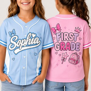 May include: A light blue baseball jersey with white trim and the name Sophia printed on the front, paired with a pink baseball jersey featuring the text First Grade, a crown, a bow, a star wand, and a backpack graphic on the back.