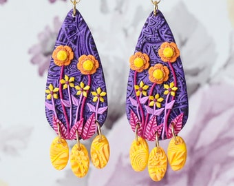 Op de afbeelding: Paarse traanvormige oorbellen met gele en roze bloemmotieven en gouden accenten. De oorbellen hebben drie hangende gele ovalen.