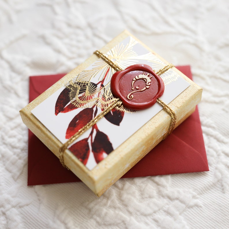 Op de afbeelding: Een klein, gouden geschenkdoosje met een rode waszegel. Het doosje is versierd met een witte achtergrond en een rood en gouden bloemmotief. De waszegel heeft een gouden ontwerp.