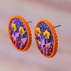 Op de afbeelding: Een paar ronde oorbellen in paars en oranje met een bloemenmotief. De oorbellen hebben een gevlochten oranje rand.