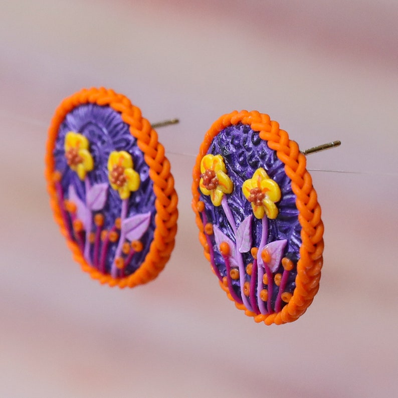 Op de afbeelding: Een paar ronde oorbellen in paars en oranje met een bloemenmotief. De oorbellen hebben een gevlochten oranje rand.