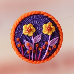 Op de afbeelding: Een ronde, oranje gerande, paarse achtergrond met twee gele bloemen en paarse bladeren. De bloemen hebben oranje centra en de bladeren hebben een zilveren glans.