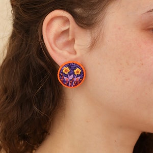 Op de afbeelding: Een paar ronde oorbellen in paars en oranje met een bloemenmotief. De oorbellen hebben kleine gele bloemen met roze stelen en bladeren.