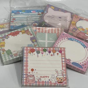 May include: A set of colorful memo pads with various designs featuring cute cartoon animals, balloons, and text. The pads are decorated with pastel colors and checkered patterns. The text on one pad reads "Happy".