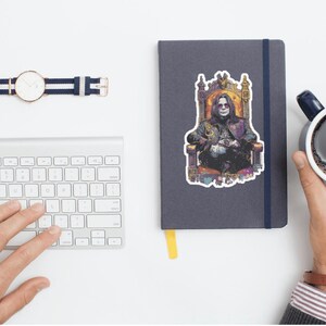 May include: A flat lay featuring a silver keyboard, a navy blue notebook with a sticker of a person on a throne, a watch with a navy blue and white band, and a mug of coffee. A hand is typing on the keyboard.