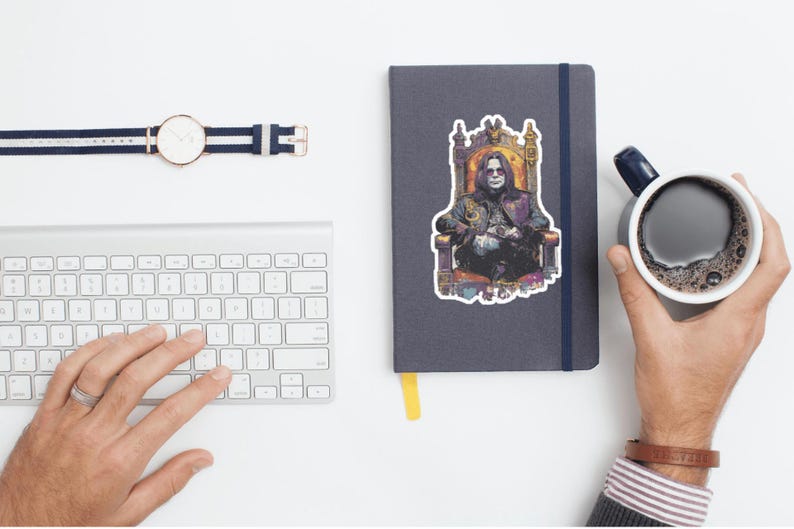May include: A flat lay featuring a silver keyboard, a navy blue notebook with a sticker of a person on a throne, a watch with a navy blue and white band, and a mug of coffee. A hand is typing on the keyboard.