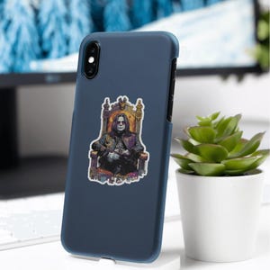 May include: A navy blue phone case with a sticker featuring a seated figure in a throne. The sticker has a dark color palette with gold and purple accents. The phone case is displayed on a white stand, with a small potted plant to the right.