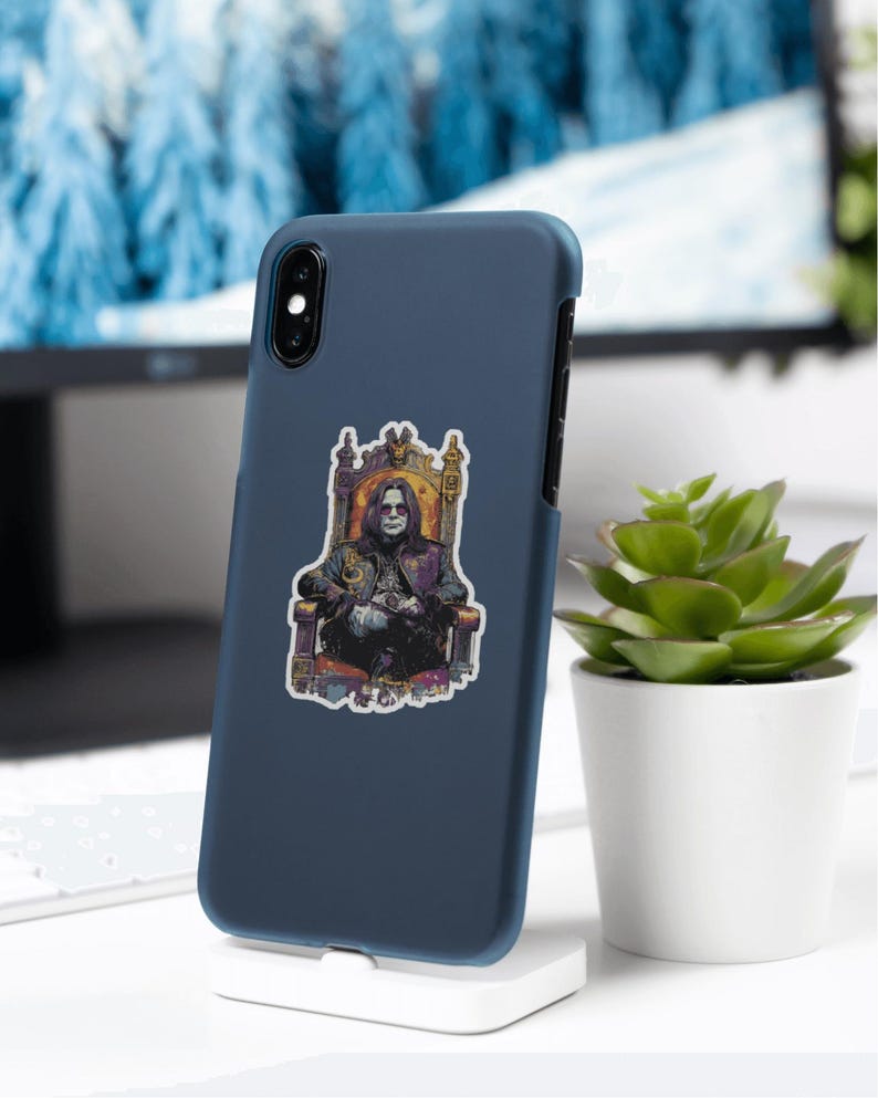 May include: A navy blue phone case with a sticker featuring a seated figure in a throne. The sticker has a dark color palette with gold and purple accents. The phone case is displayed on a white stand, with a small potted plant to the right.