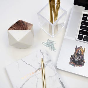 May include: A white and wood-toned geometric container, a clear acrylic pen holder with gold pens, a marble notebook, a gold pen, paper clips, and a laptop with a sticker of a person on a throne. The items are arranged on a white surface.