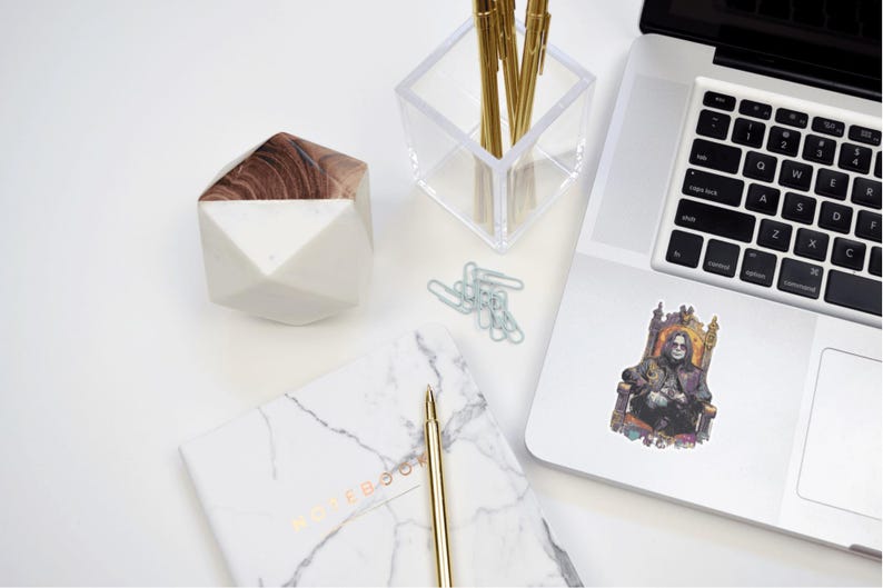 May include: A white and wood-toned geometric container, a clear acrylic pen holder with gold pens, a marble notebook, a gold pen, paper clips, and a laptop with a sticker of a person on a throne. The items are arranged on a white surface.