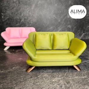 Könnte beinhalten: Ein grünes Mini-Sofa mit zwei Kissen und Holzbeinen. Das Sofa steht auf einer grauen Oberfläche. Der Text "ALIMA UNIQUE" ist in der oberen rechten Ecke des Bildes sichtbar.
