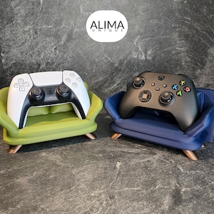 Könnte beinhalten: Zwei Miniatur-Sofas, eines grün und eines blau, jedes mit einem Videospiel-Controller. Das grüne Sofa hält einen weißen PlayStation 5-Controller, während das blaue Sofa einen schwarzen Xbox-Controller hält.