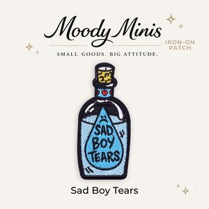 May include: An embroidered iron-on patch shaped like a glass bottle with a cork stopper. The bottle contains light blue liquid with a central teardrop graphic featuring a sad face and the text SAD BOY TEARS in black lettering. The neck has red hearts.