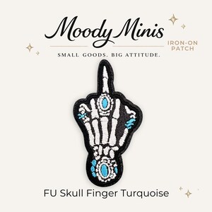 May include: An iron-on patch featuring an embroidered skeleton hand making a middle finger gesture. The design includes white bones, black background, and bright turquoise accents on the rings and knuckles. Text reads Moody Minis, Iron-On Patch, Small Goods. Big Attitude. FU Skull Finger Turquoise.