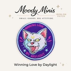 May include: A circular embroidered iron-on patch featuring a white cat with yellow eyes and a crescent moon on its forehead. The cat is snarling, surrounded by pink lightning bolts and blue stars on a purple background with the text Winning Love by Daylight.