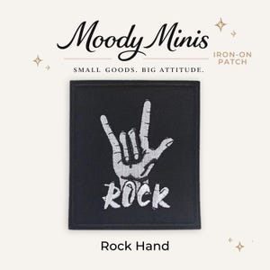 May include: A rectangular black iron-on patch featuring a white embroidered graphic of a hand making the rock and roll sign. Below the hand, the word ROCK is embroidered in white capital letters. The patch has a clean, stitched border.